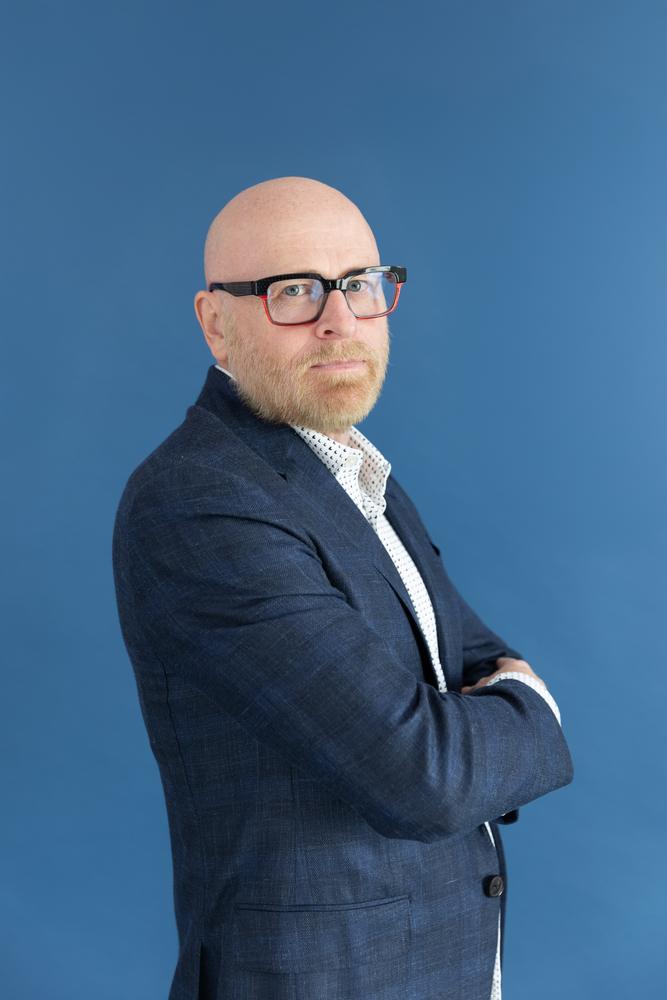 Portrait of a man in a navy blazer with arms crossed on a blue backdrop