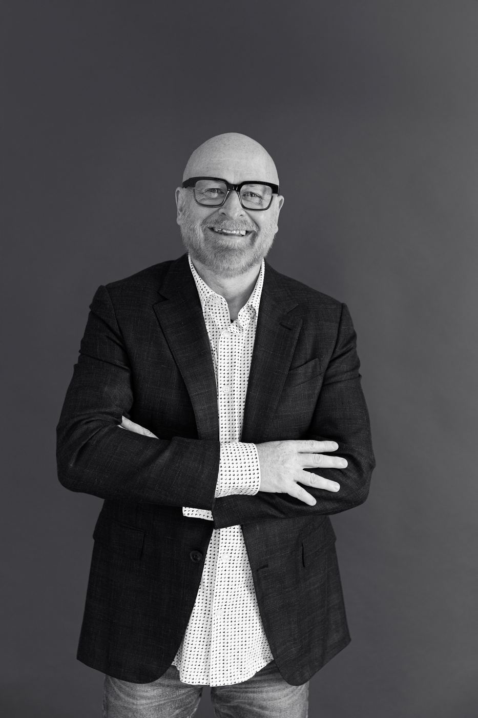 Black and white headshot of a laughing man in a patterned shirt and blazer with arms crossed
