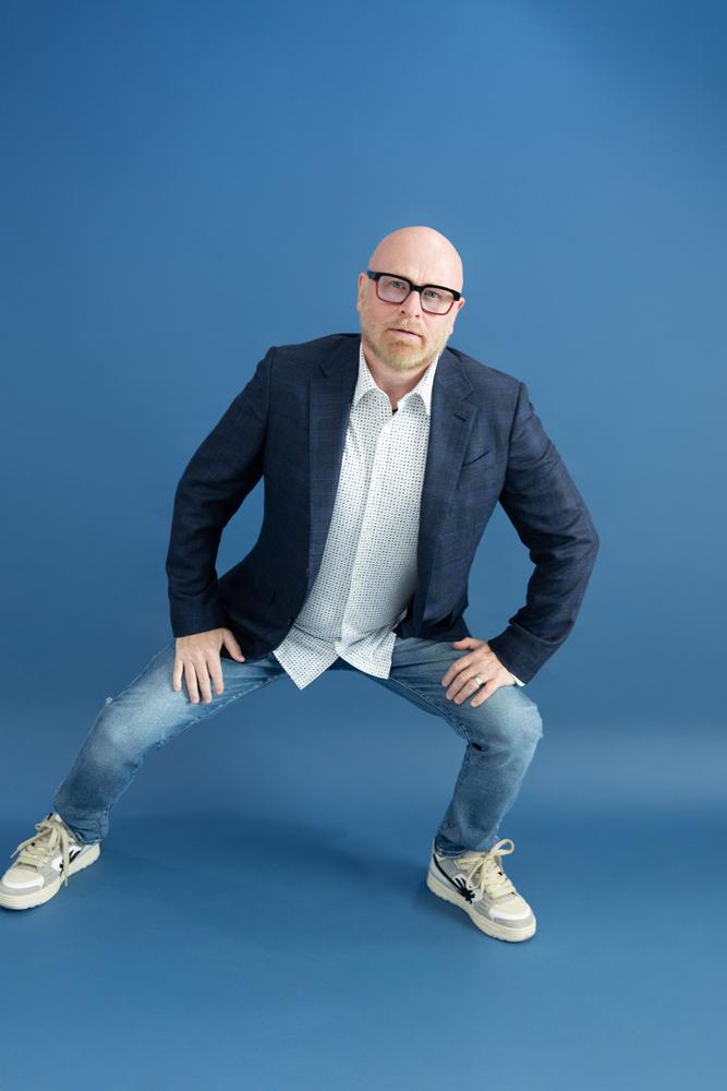 Playful full-length portrait of a man in a blazer and jeans crouching on a blue backdrop