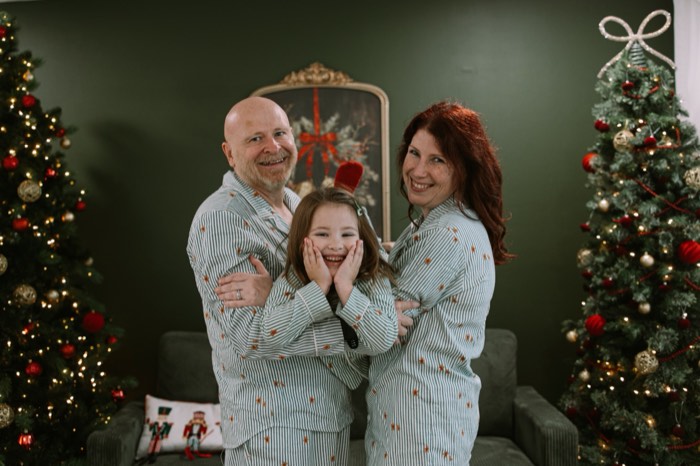 Mom, dad and daughter in matching holiday pajamas between the studio Christmas trees